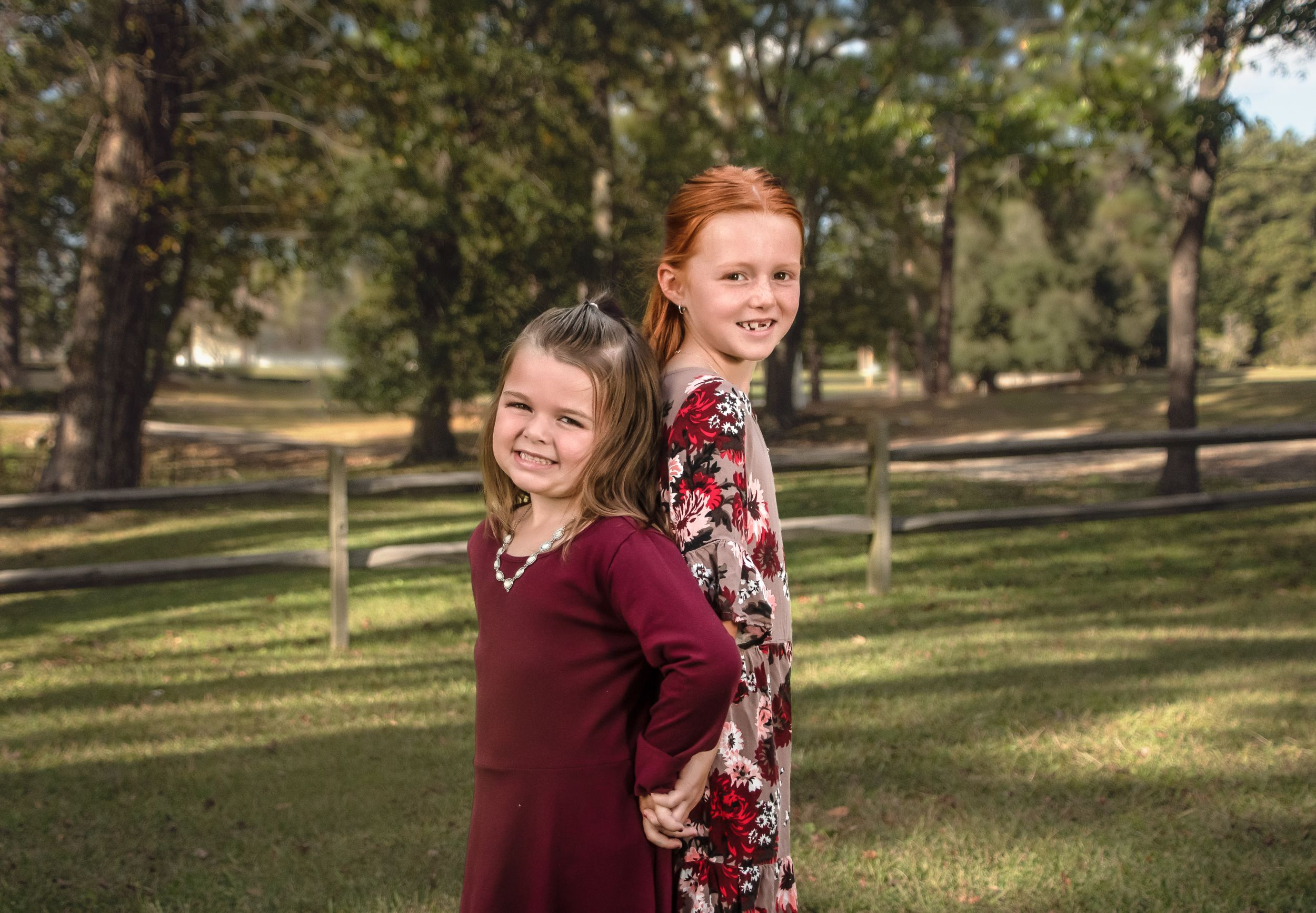 Outdoor portrait of two girls in burgundy dresses posing back-to-back in a park setting.