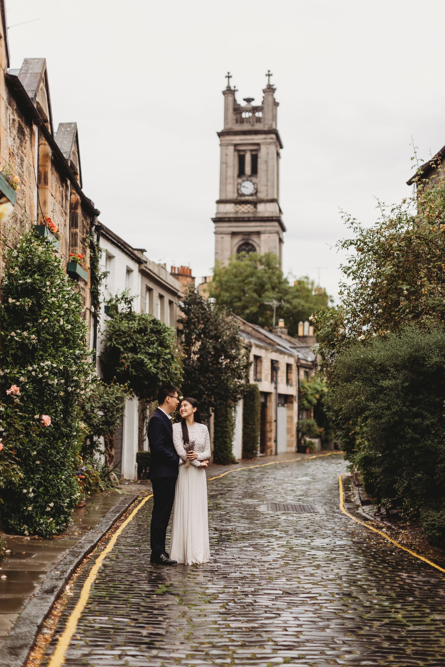 A couple in formal wear embraces on a rainy cobblestone street with church spire in background.