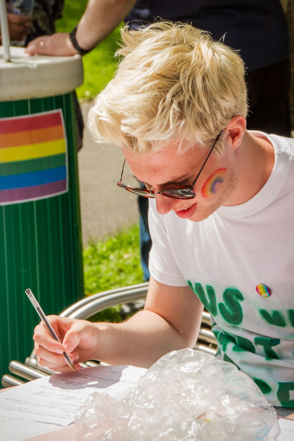 Person with colorful face paint signing documents at Pride event.