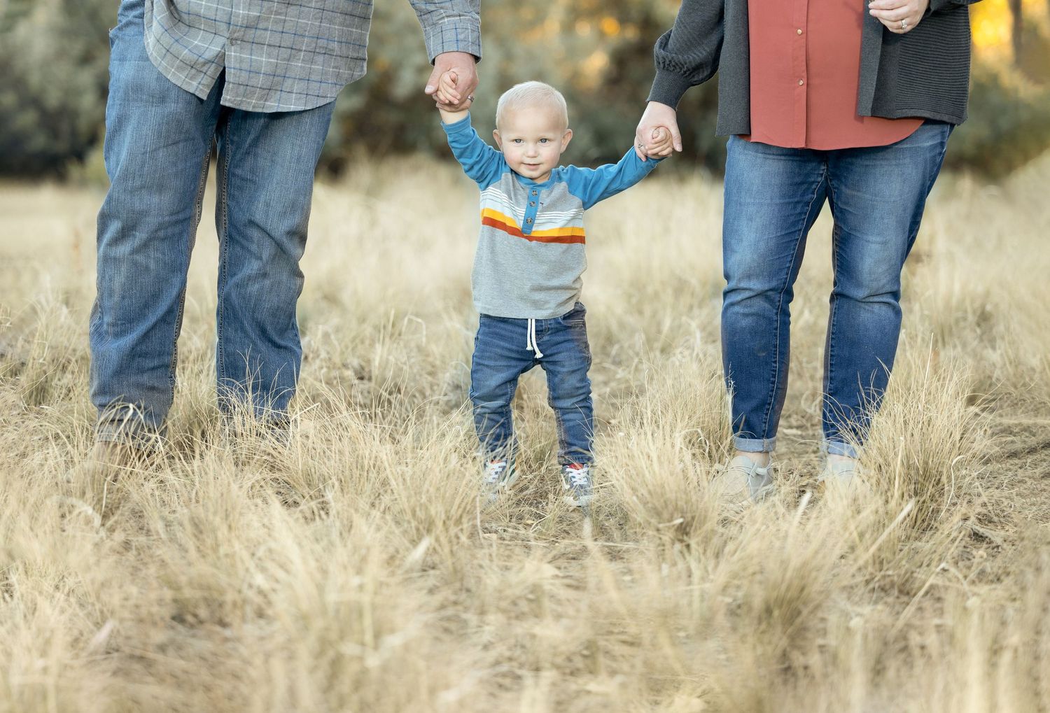 Small child taking first steps in tall grass field while holding hands with adults.