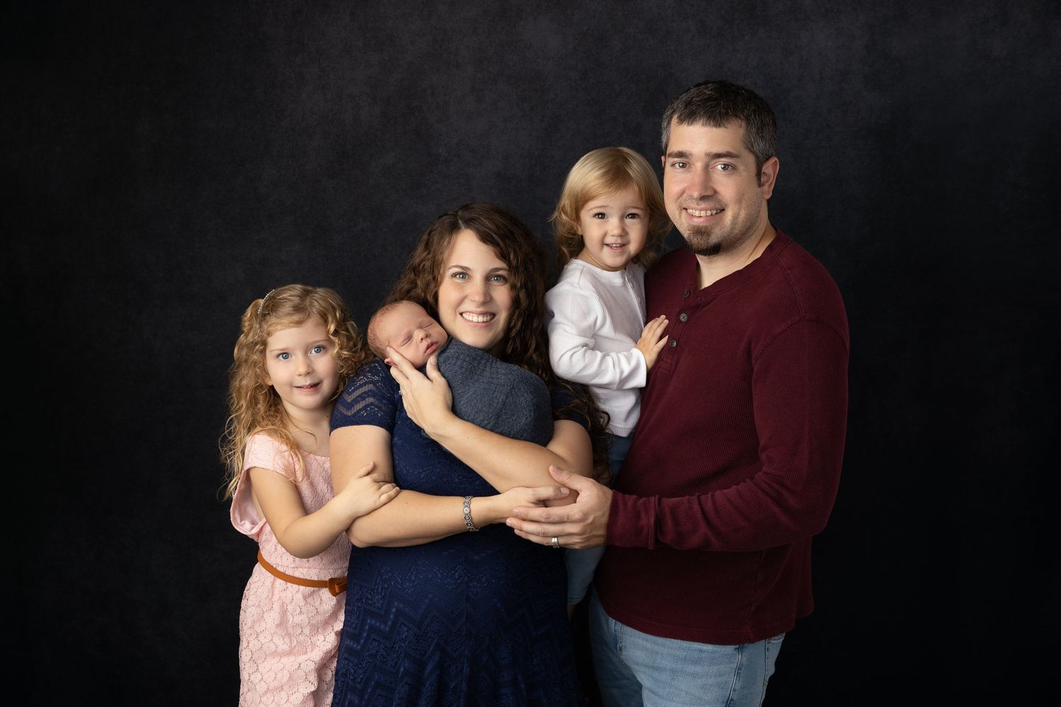 Family group poses informally together while wearing coordinating outfits against a dark backdrop.