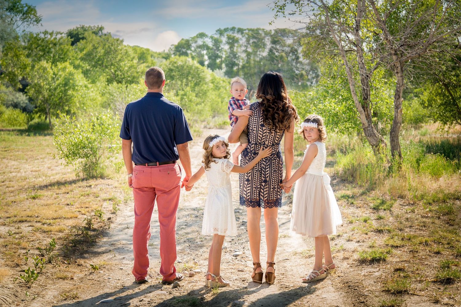 Family walks together holding hands on a sunlit nature path surrounded by green trees.