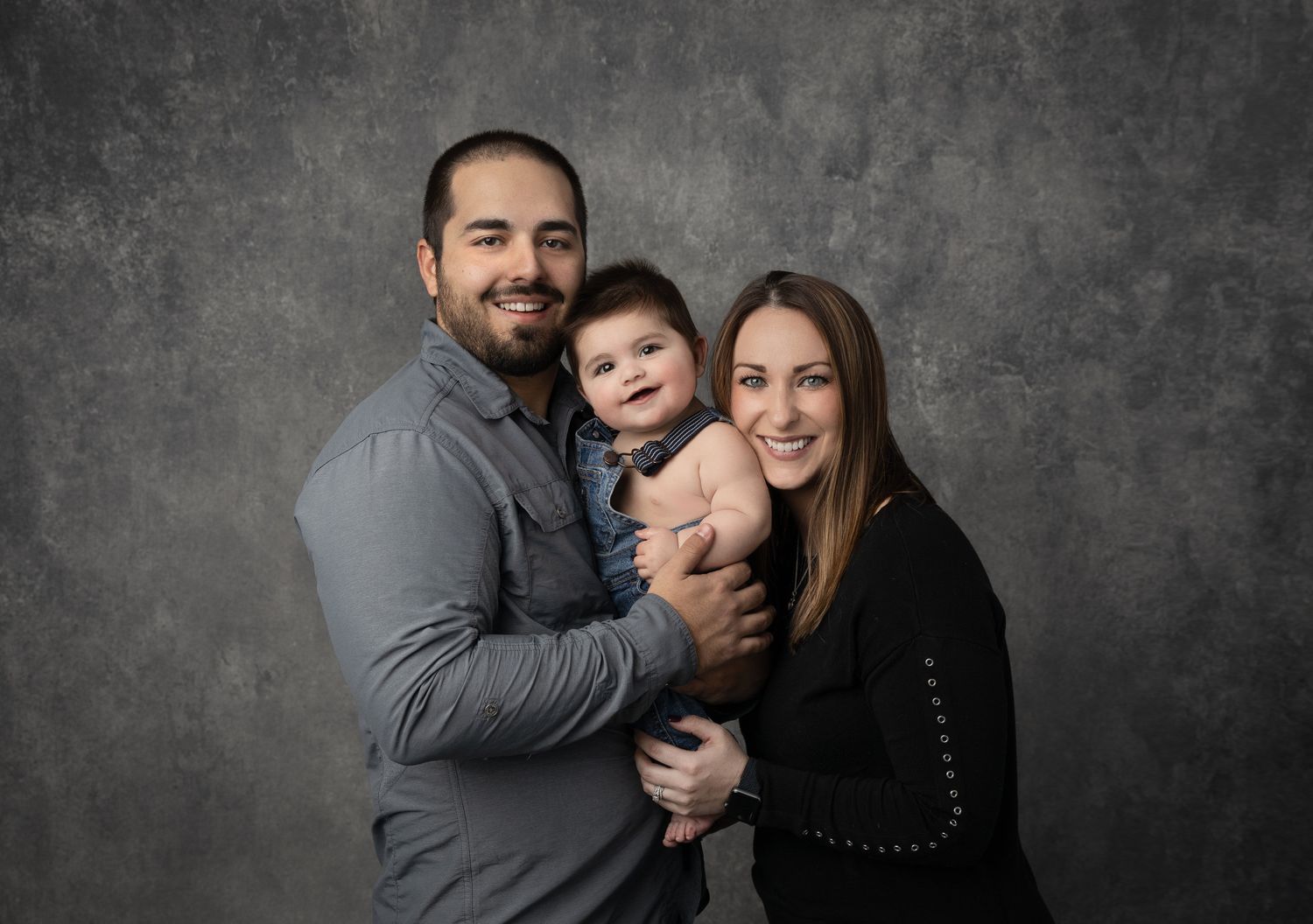 Intimate family portrait against textured grey background shows close embraces and warm smiles.