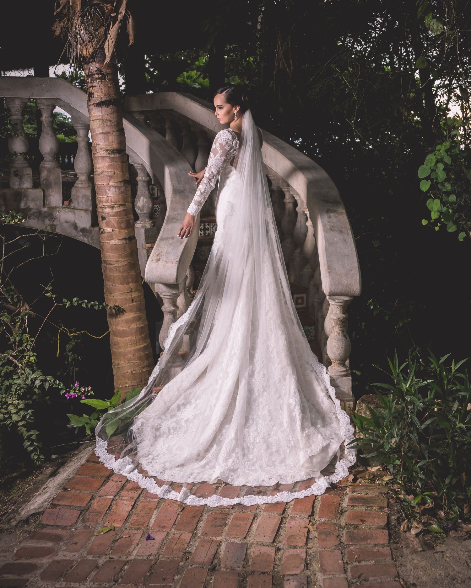 Dramatic bridal portrait on brick path with flowing lace train and garden backdrop.