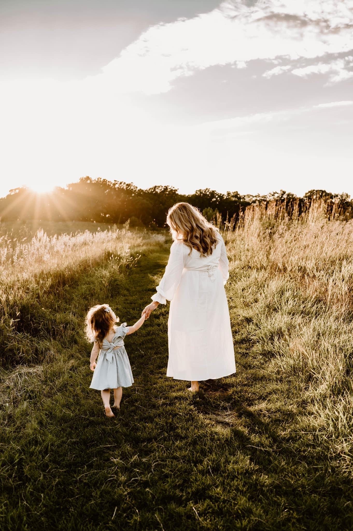 A mother and child hold hands while walking through a sunlit field at golden hour.