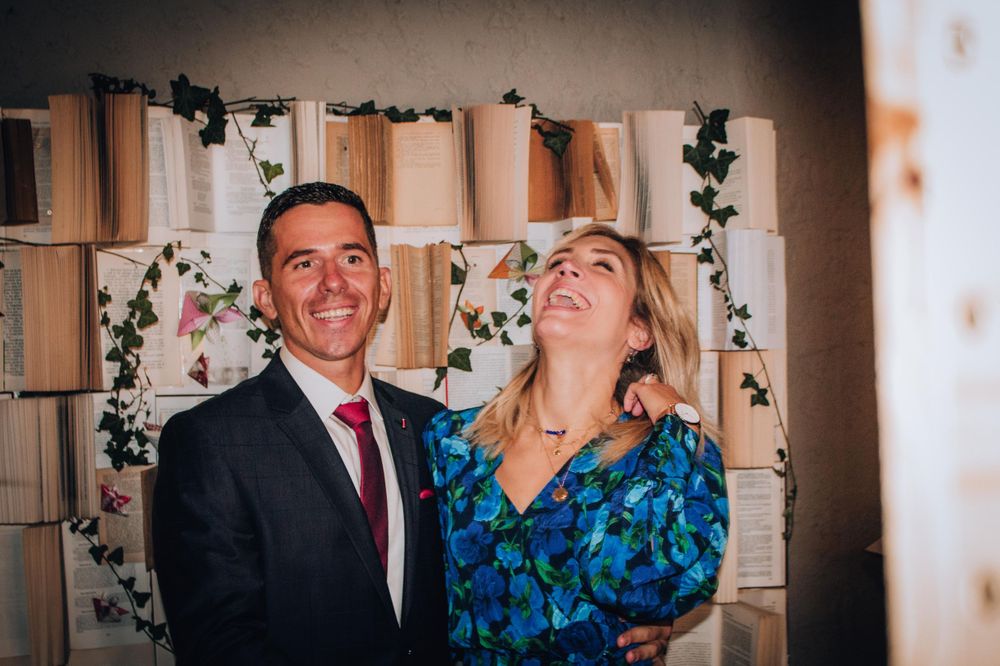 Couple laughing together against backdrop of books and greenery at wedding venue.