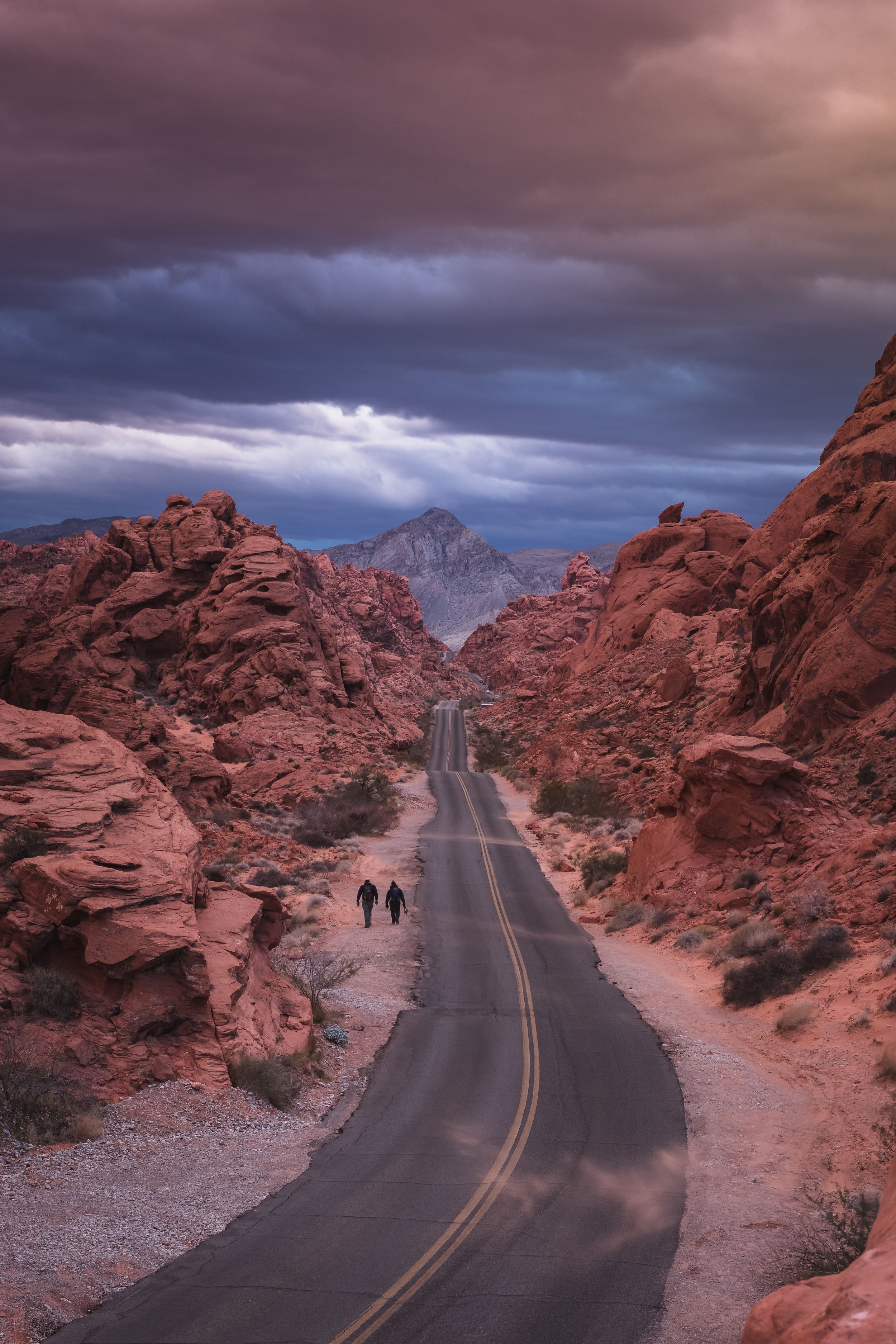 Two hikers walking down empty Mouse Tank Road in Valley of Fire State Park during sunset.