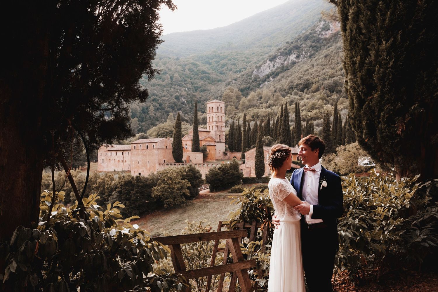 A couple embraces in formal attire against a scenic mountainside backdrop with ancient architecture in the distance.