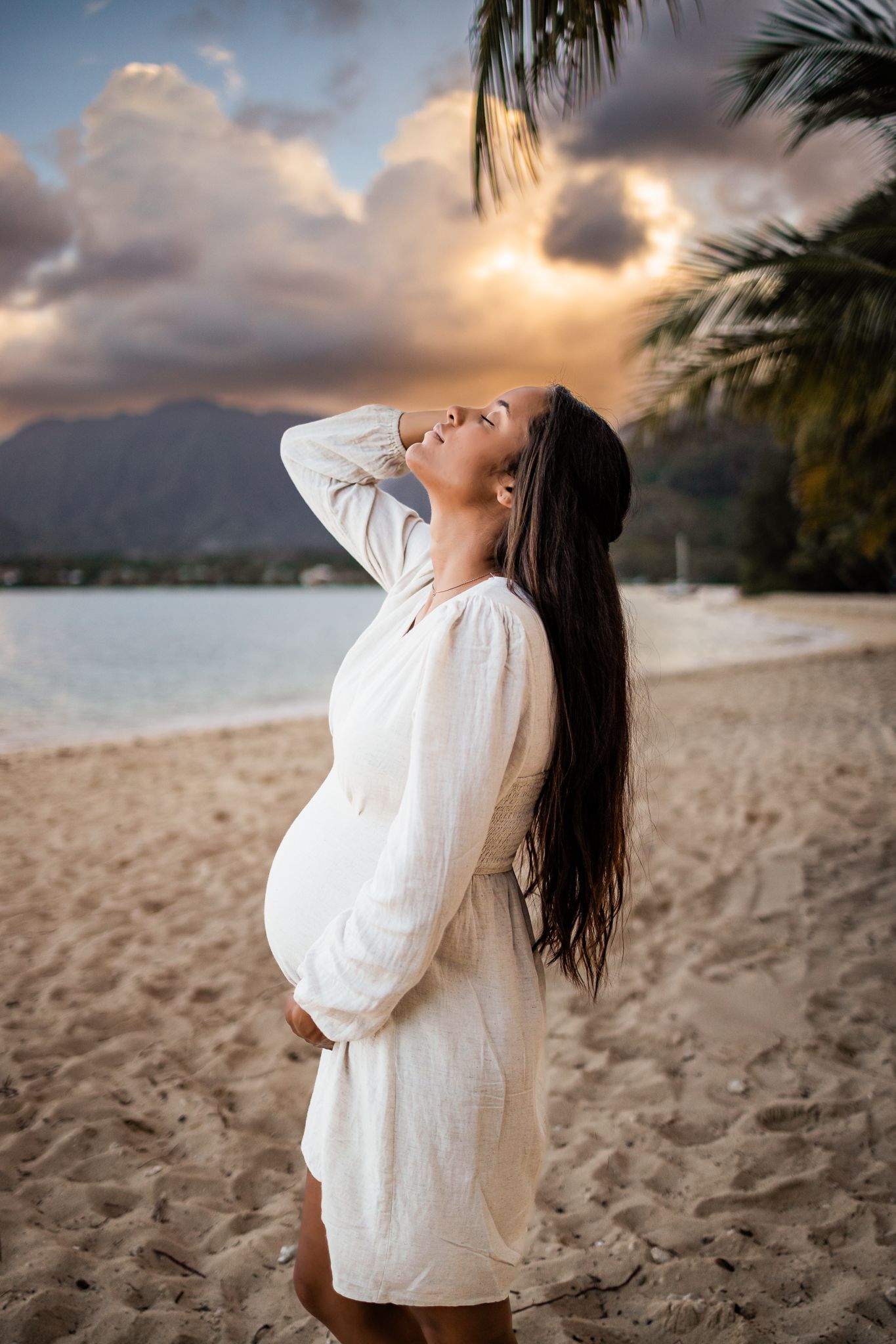 Woman in white dress poses on beach at sunset with mountains and palm trees in background.