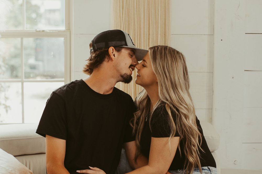 Intimate couple shares tender moments on white couch in sunlit room during romantic photoshoot.
