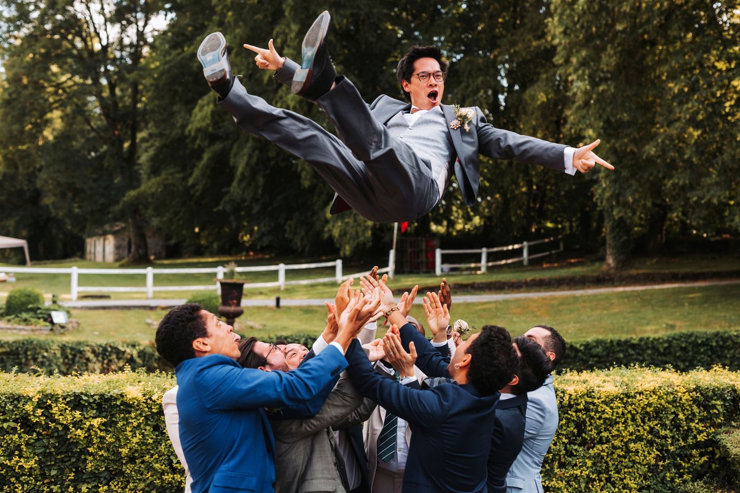 Groomsmen in blue suits toss someone horizontally in the air near a manicured garden hedge.