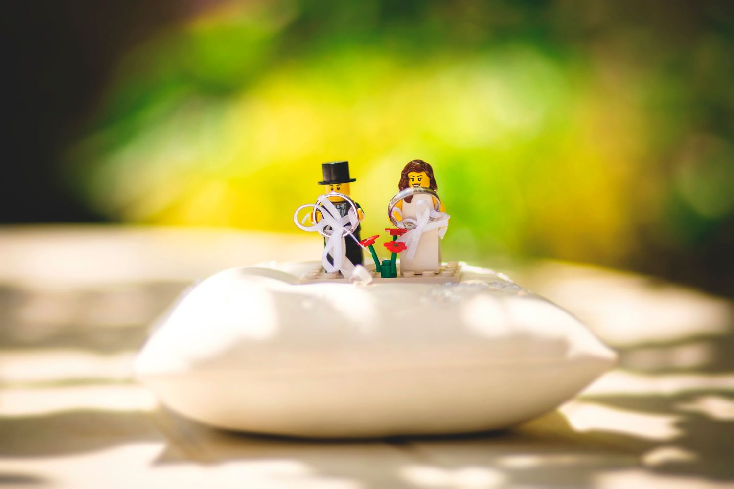 Miniature bride and groom cake toppers positioned on white cushion with blurred background.