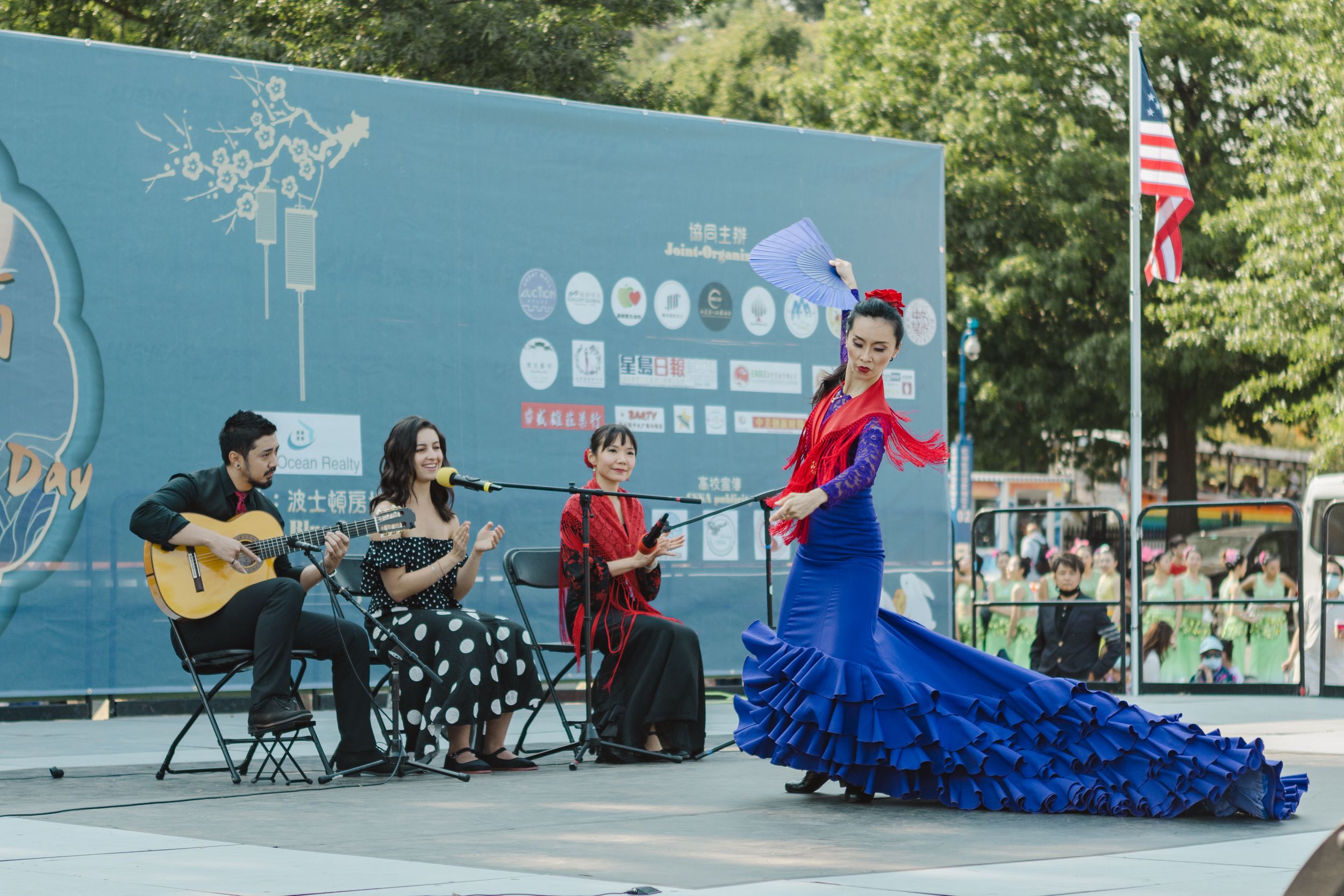 Performers in traditional costumes on an outdoor stage with American flag in background.