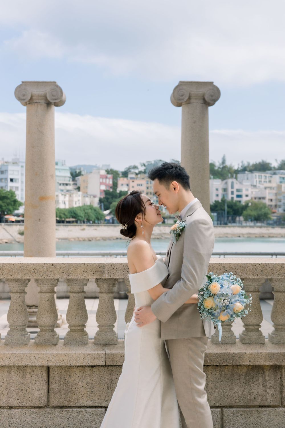 A couple in wedding attire embraces on a waterfront terrace with stone balustrades.