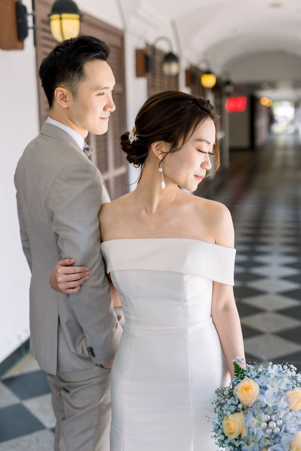 A wedding couple in gray suit and white dress embrace while holding a blue and yellow floral bouquet.