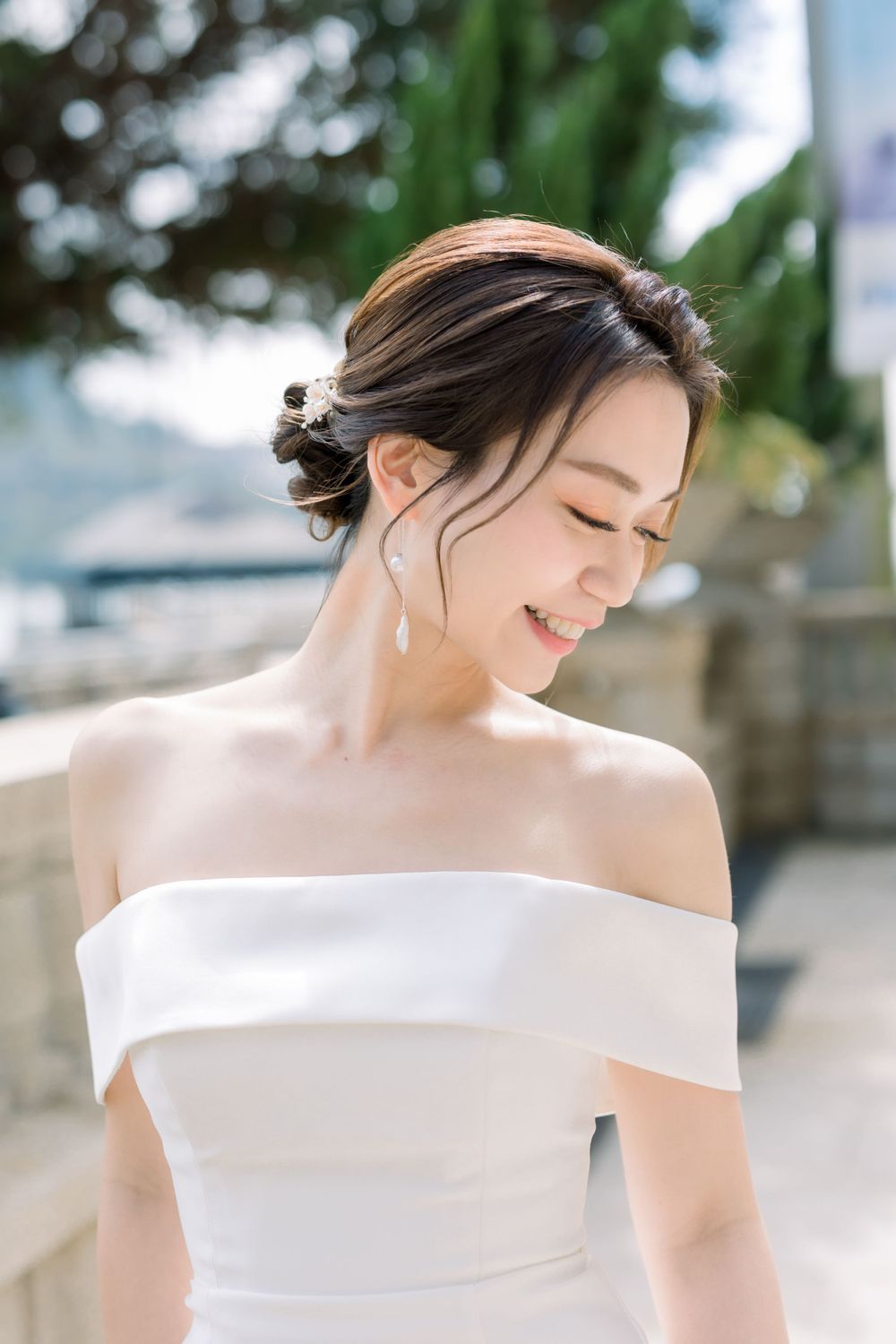 Elegant bride in off-shoulder white dress smiles with natural sunlight illuminating her face.