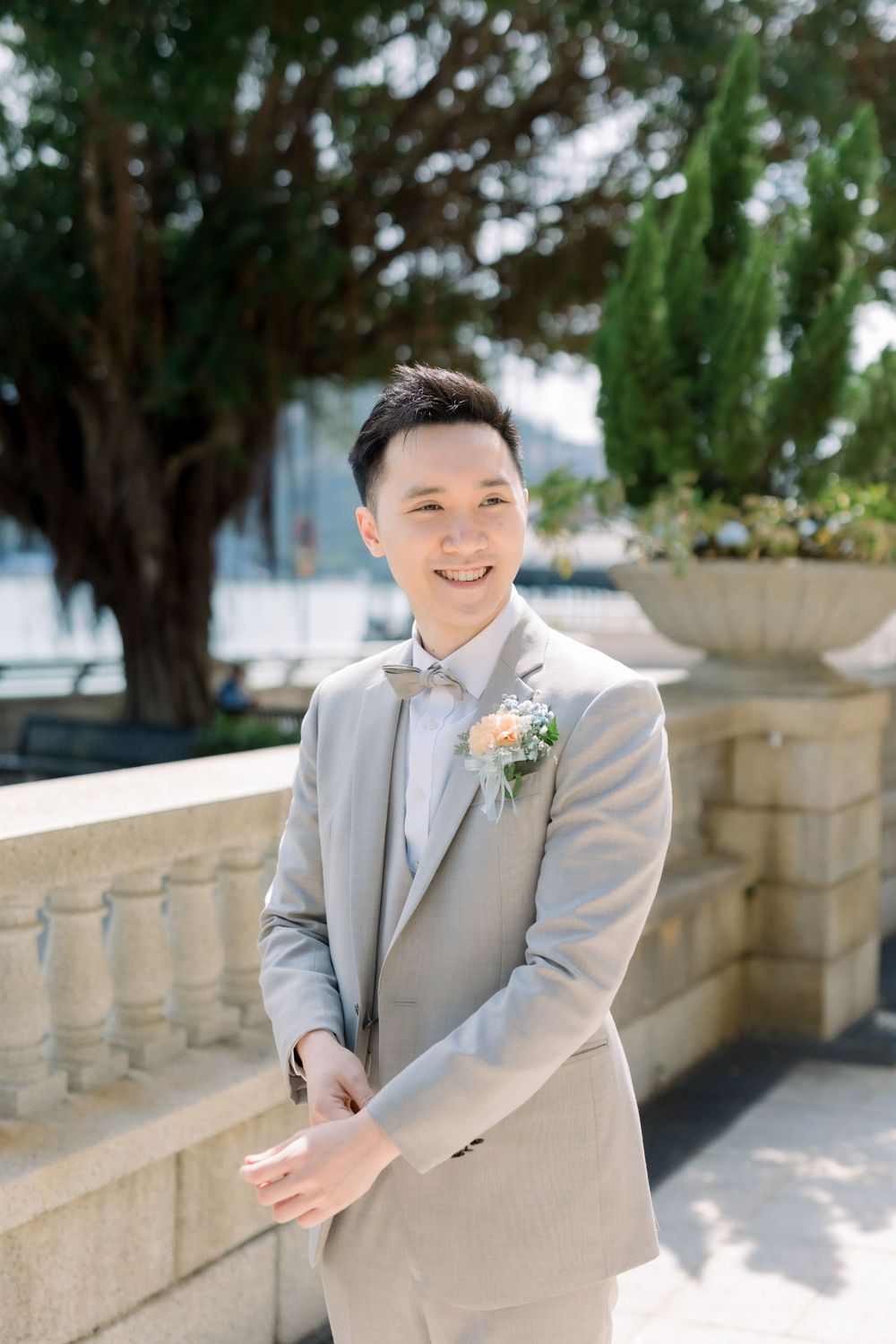 Groom in light grey suit with boutonniere smiles on stone terrace with trees in background.