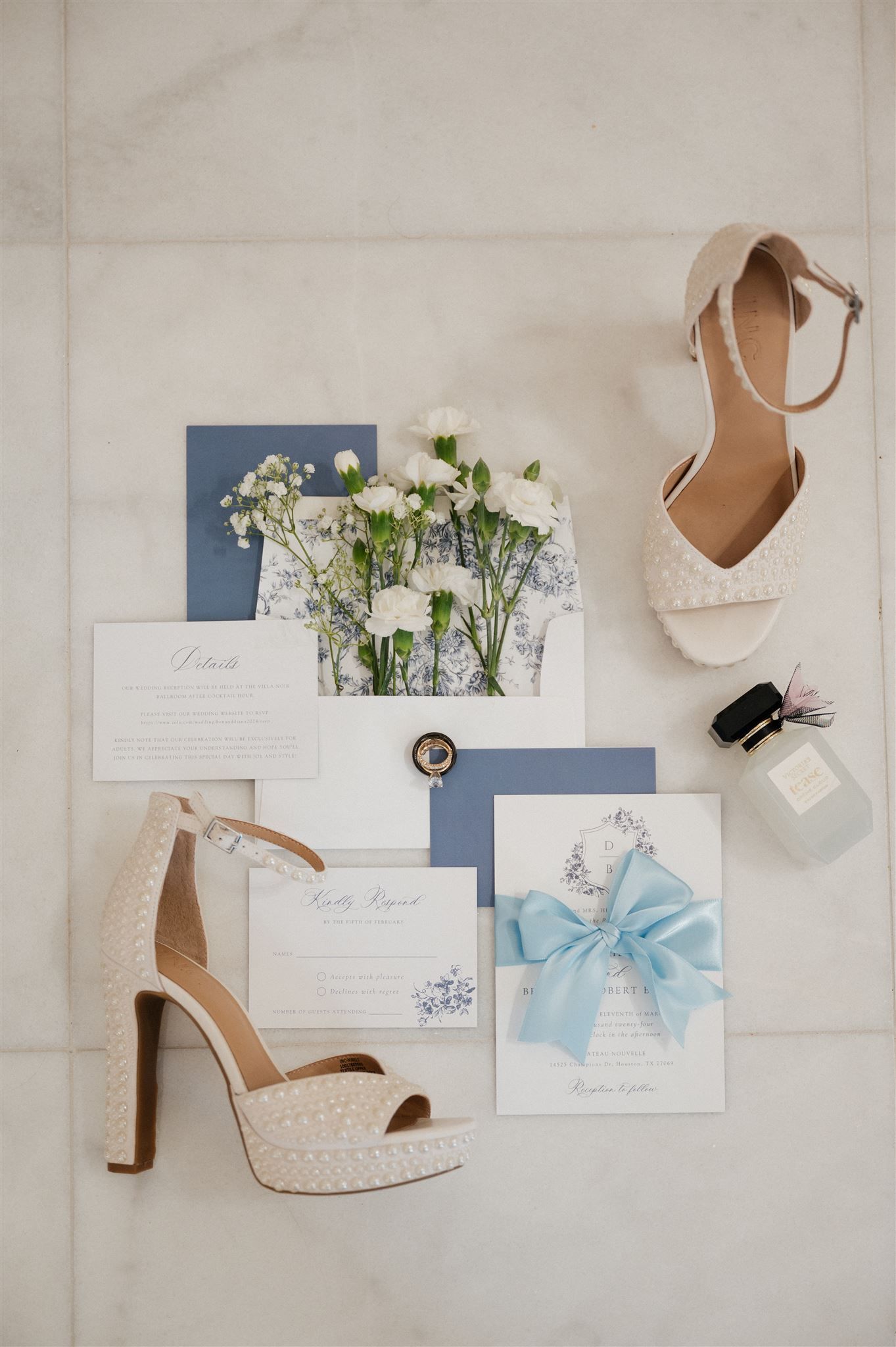 Wedding details flatlay with white peep-toe heels, floral arrangements, navy blue invitations and light blue ribbon on marble surface.