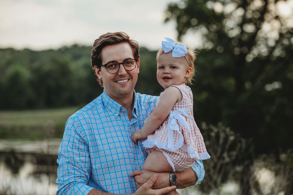 Parent holding baby against sunset backdrop during outdoor family photoshoot.