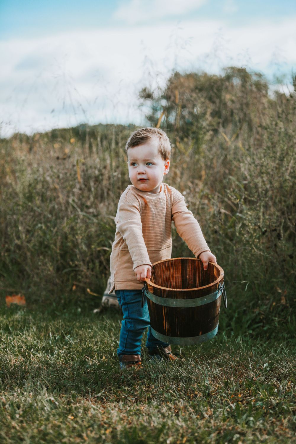 Toddler exploring outdoors with wooden basket in grassy field.