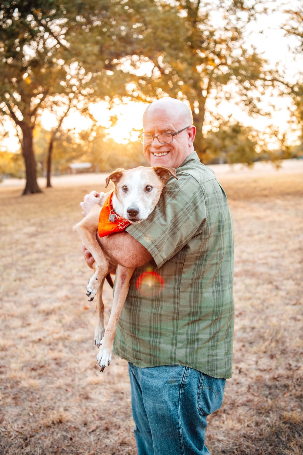 Person in green plaid shirt holds smiling dog wearing orange bandana against sunset backdrop.