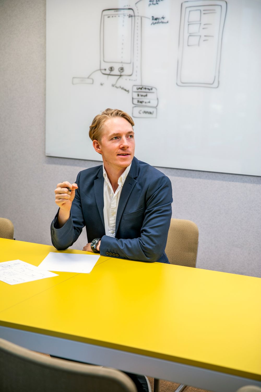 Orange County photographer captures businessman in navy blazer gestures during meeting at yellow table with whiteboard sketches behind him.