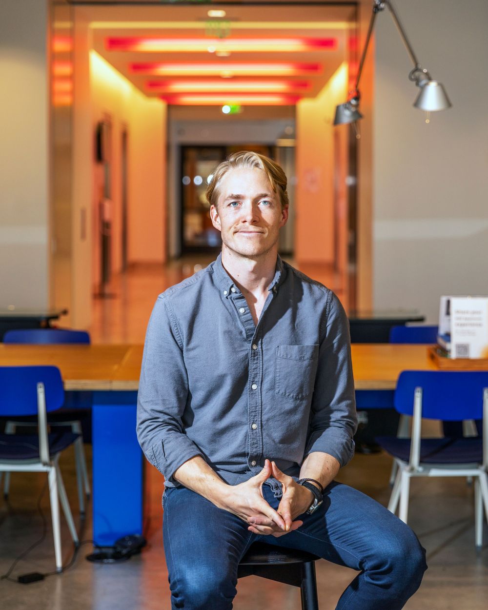 Orange County photographer captures Young man in gray shirt sits relaxed on stool in modern office lobby with colorful lighting.