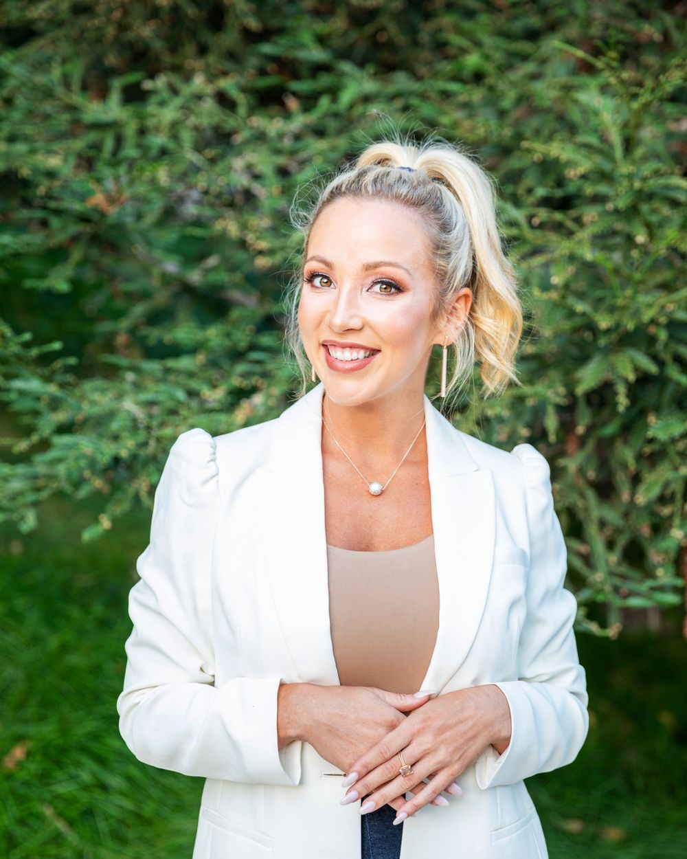 Orange County photographer captures Smiling blonde woman in white blazer posing outdoors with green trees in background.