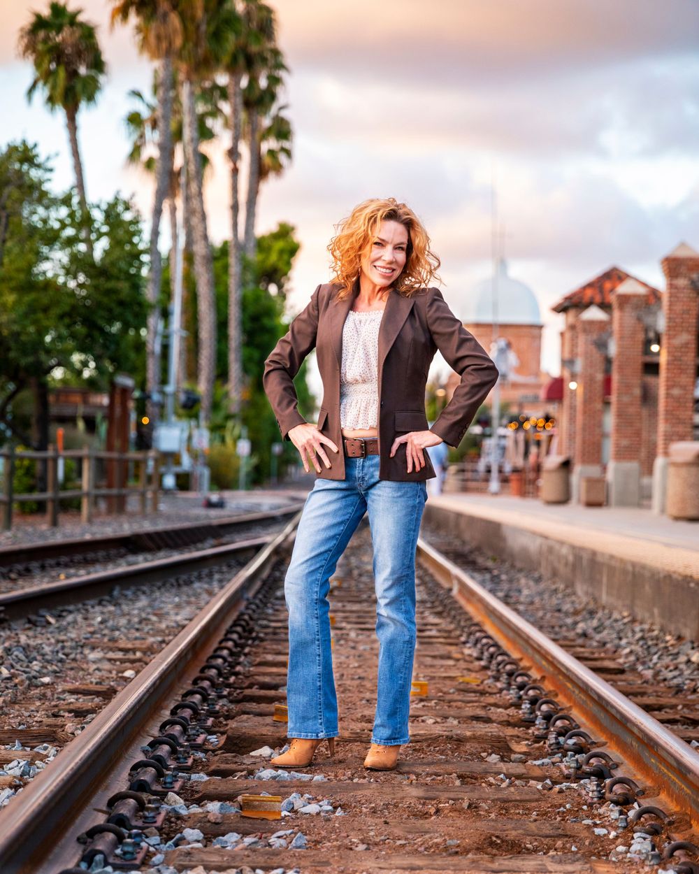 Orange County photographer captures Curly-haired woman in blazer and jeans standing on railroad tracks with palm trees at sunset.