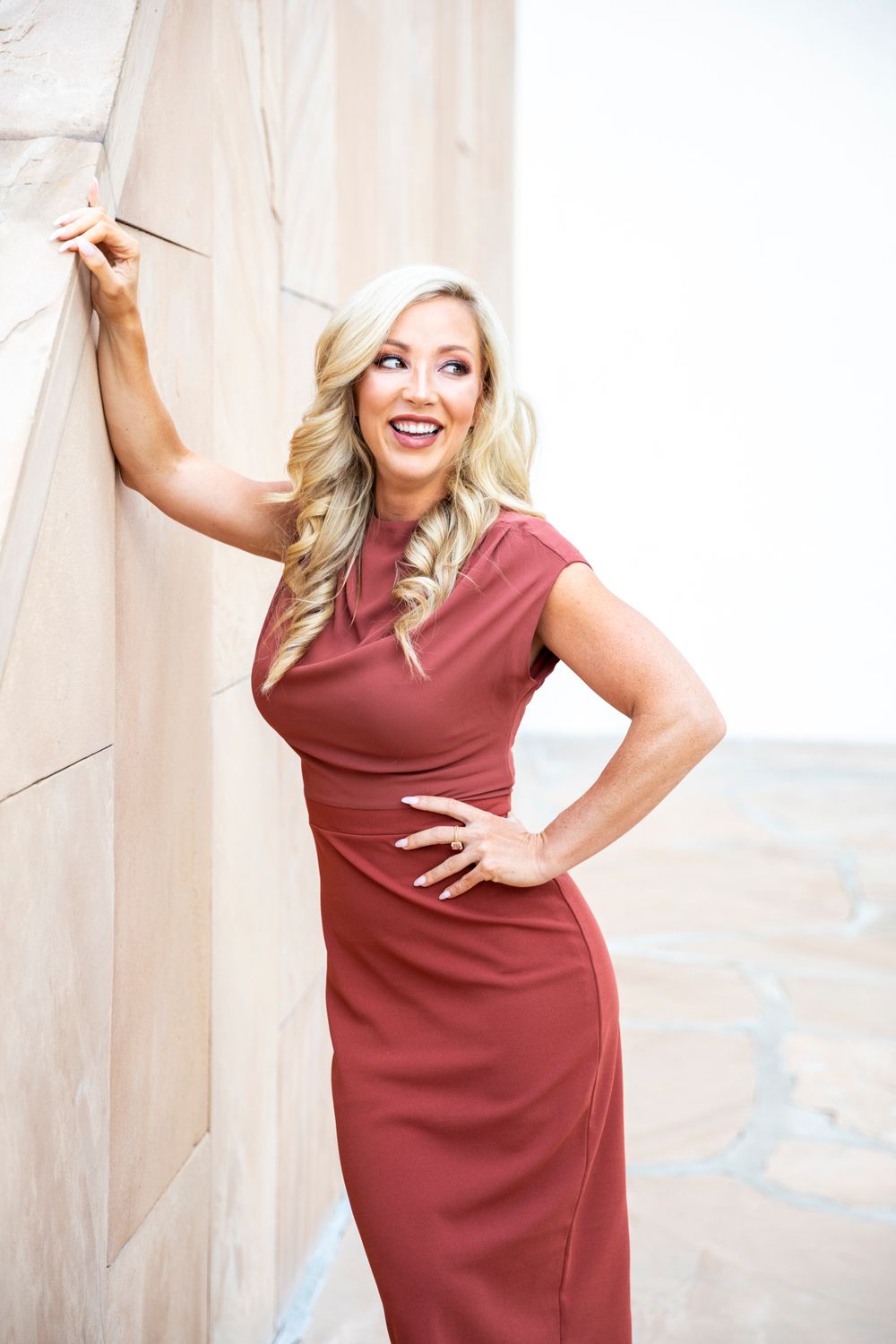 Orange County photographer captures  woman in elegant rust-colored dress poses confidently against light stone wall outdoors.