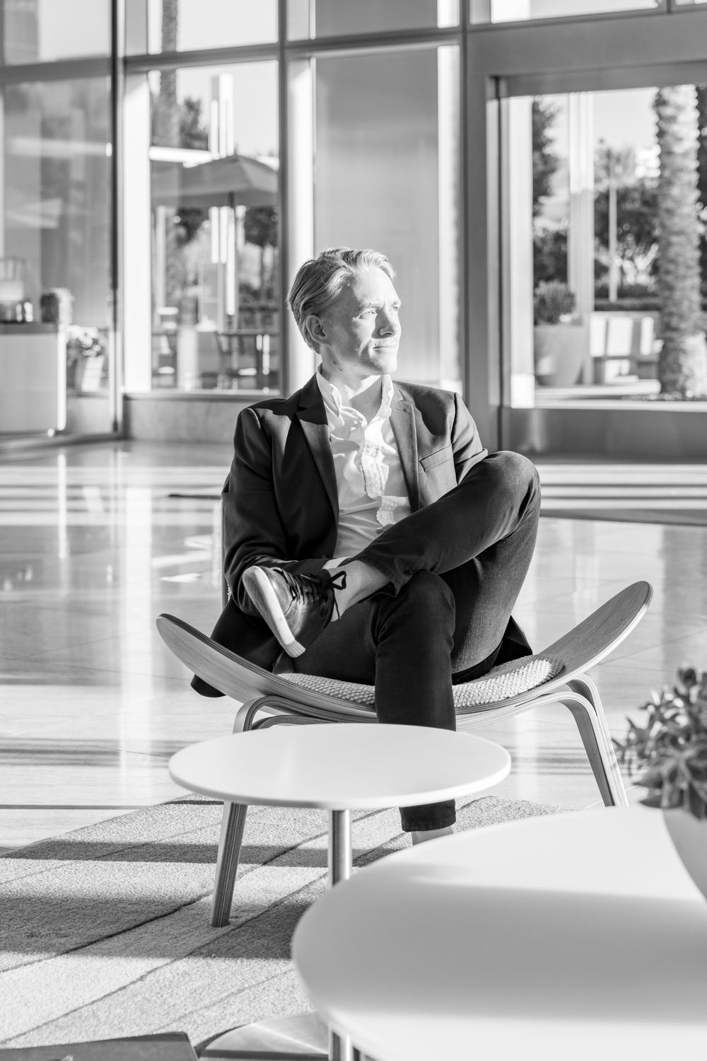 Orange County photographer captures Black and white portrait of woman sitting cross-legged in modern lobby chair near large windows.