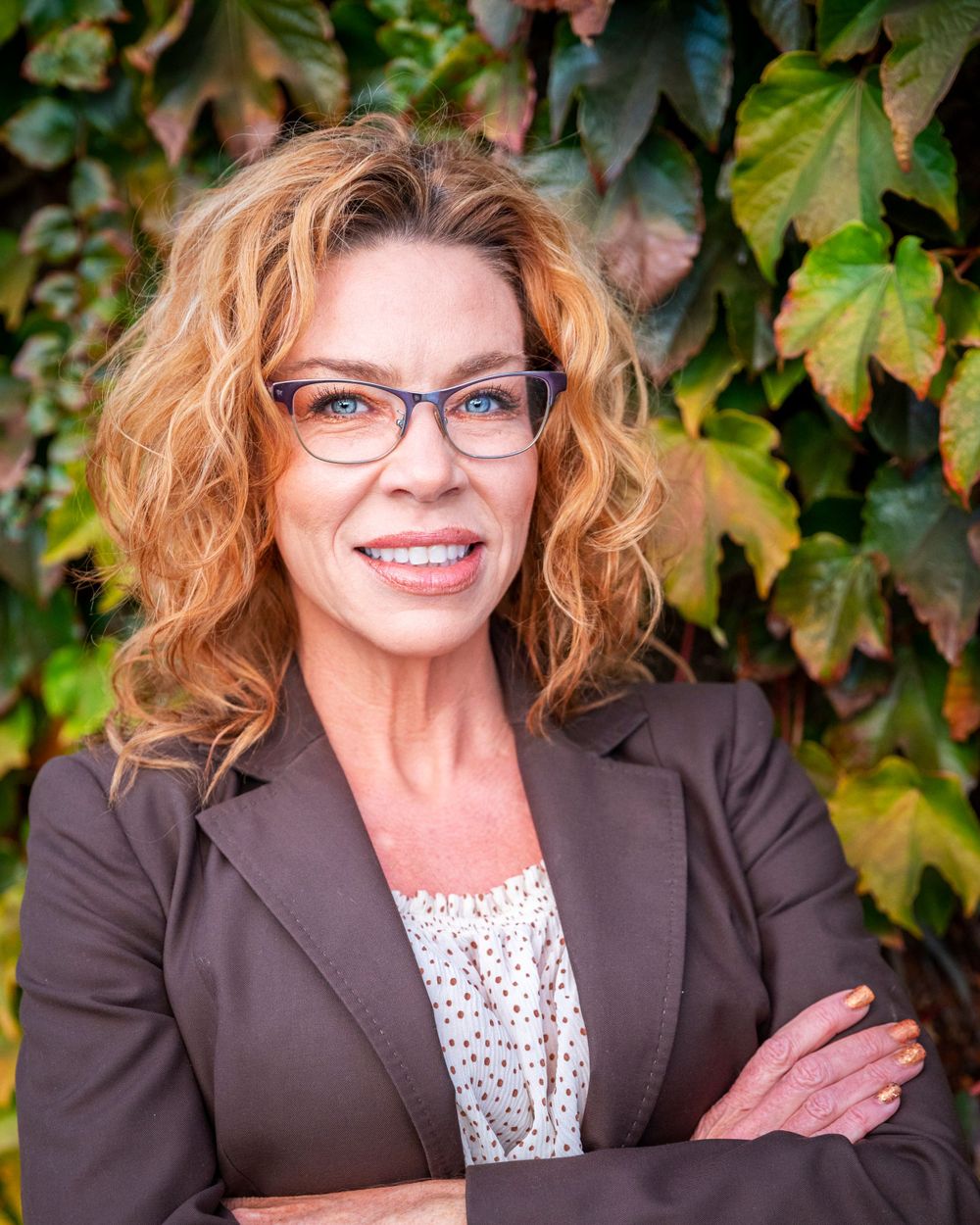Orange County photographer captures Professional woman with curly red hair and glasses smiling confidently in front of green ivy wall.