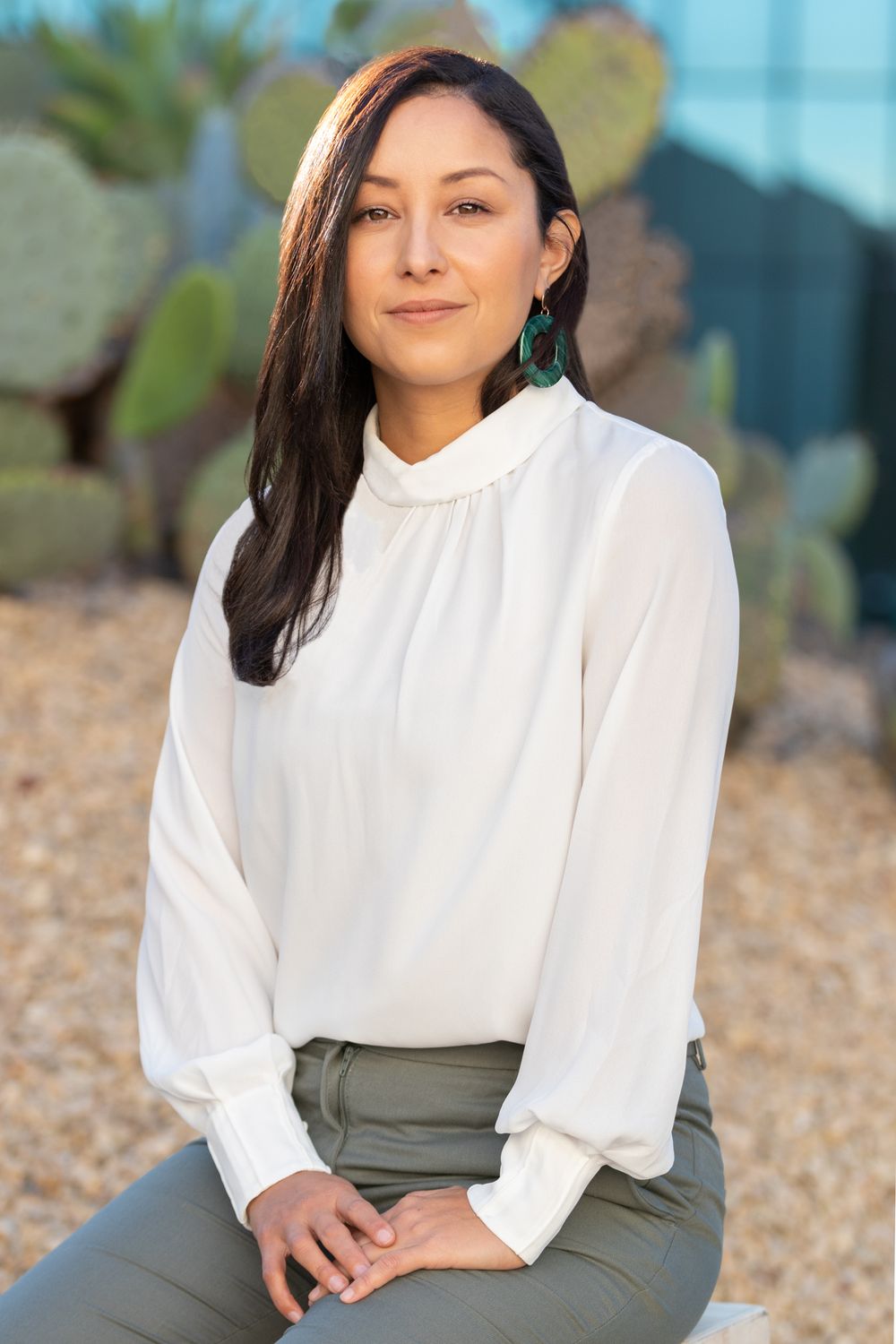 Orange County photographer captures Young woman in white blouse and green earrings sits outdoors near cacti in a professional portrait.