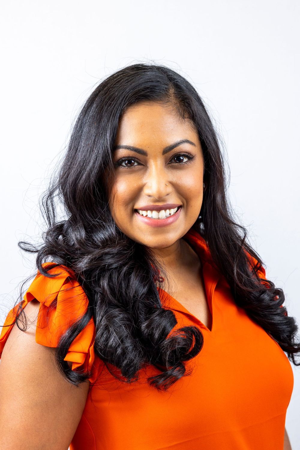 Orange County photographer captures Smiling woman with long dark curly hair wearing an orange ruffled top against a white background.