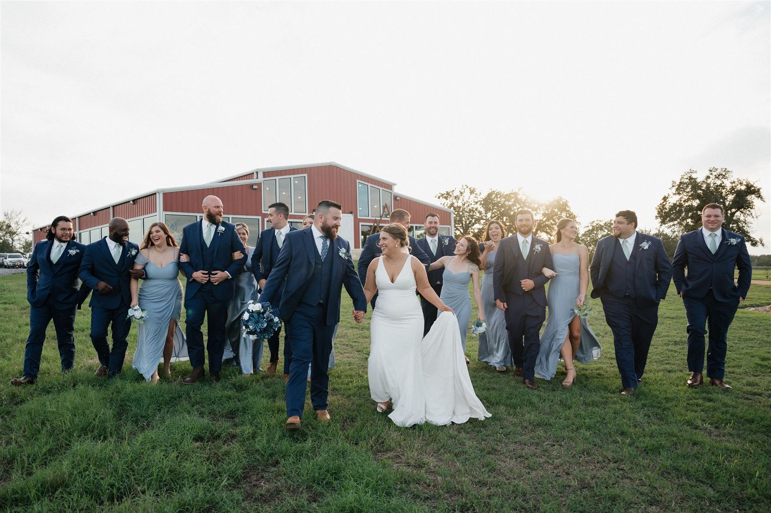 Wedding party posed together on grassy field with brick building in background.