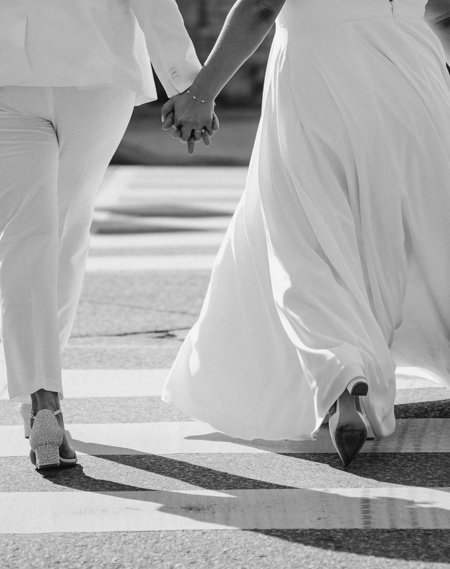 Black and white closeup of two people in white clothing holding hands while walking.