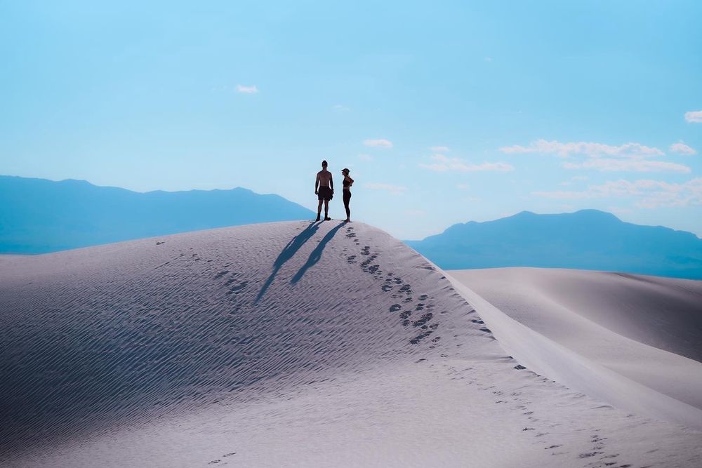 Two silhouettes stand atop a white sand dune against a backdrop of mountains and blue sky.