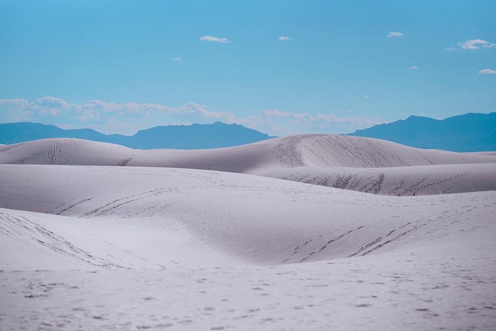 Rolling white sand dunes stretch across the landscape beneath a bright blue sky with mountains in the distance.
