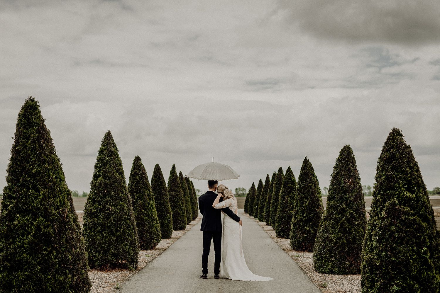 Bride and groom share a romantic moment under an umbrella along a tree-lined garden path on a cloudy wedding day.