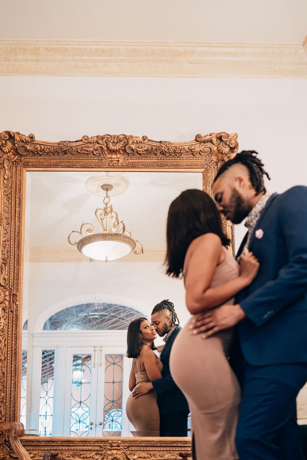 A couple's reflection appears in an ornate gold mirror as they embrace in a elegant hallway.