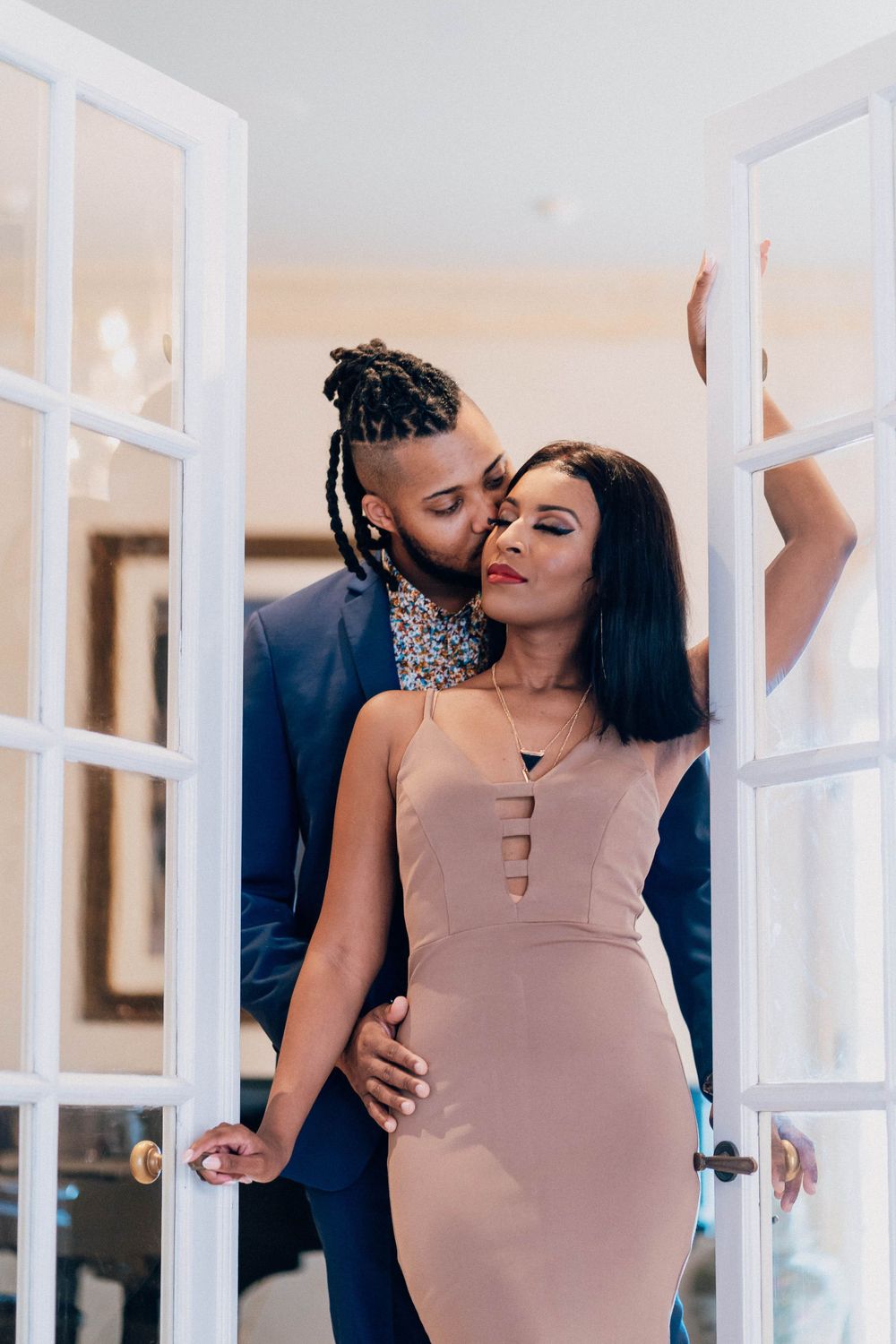 A couple shares an intimate moment between white French doors in formal attire.