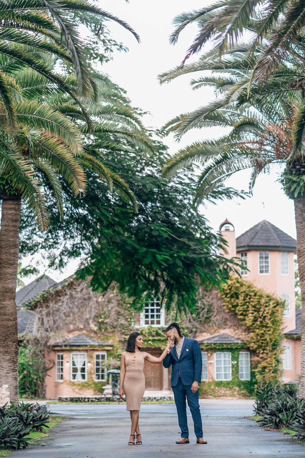 A couple walks together on a path lined with palm trees near a pink stucco building.