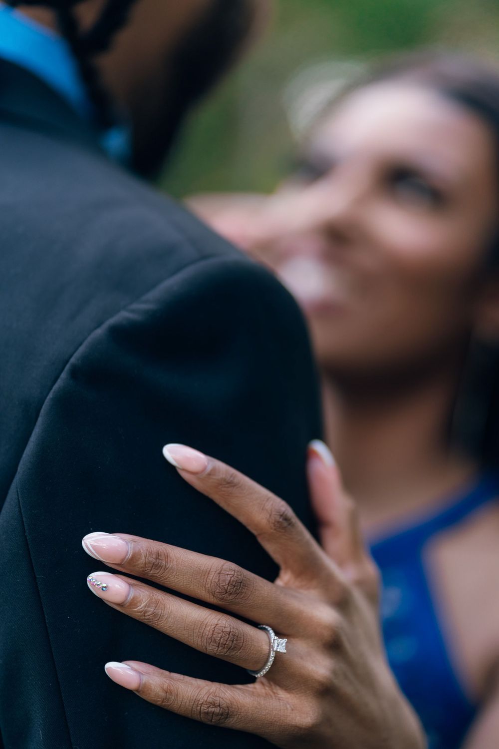 Close-up of a hand wearing an engagement ring gently resting on a black suit jacket.