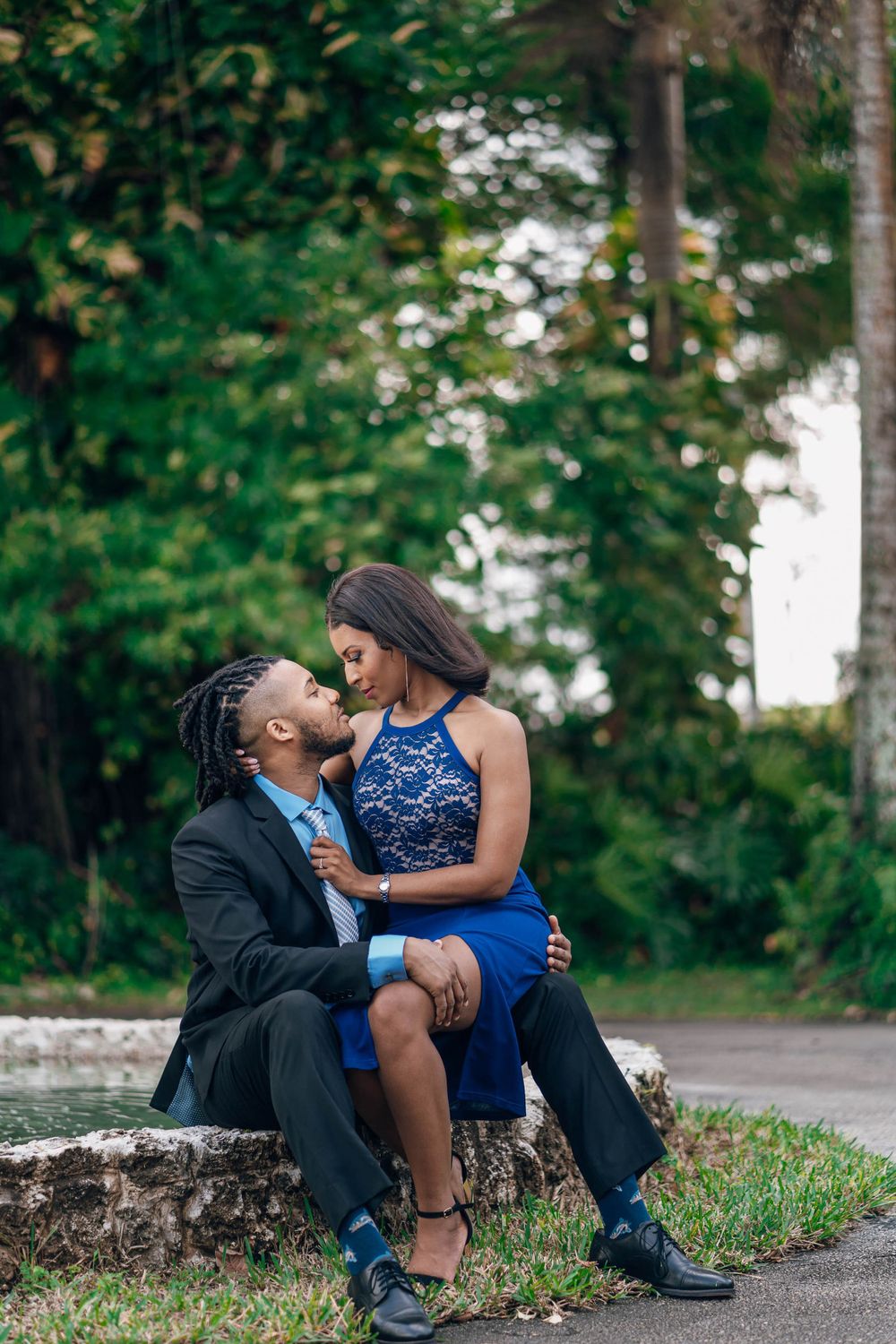 Romantic couple in formal attire sharing an intimate moment on a stone wall surrounded by greenery.