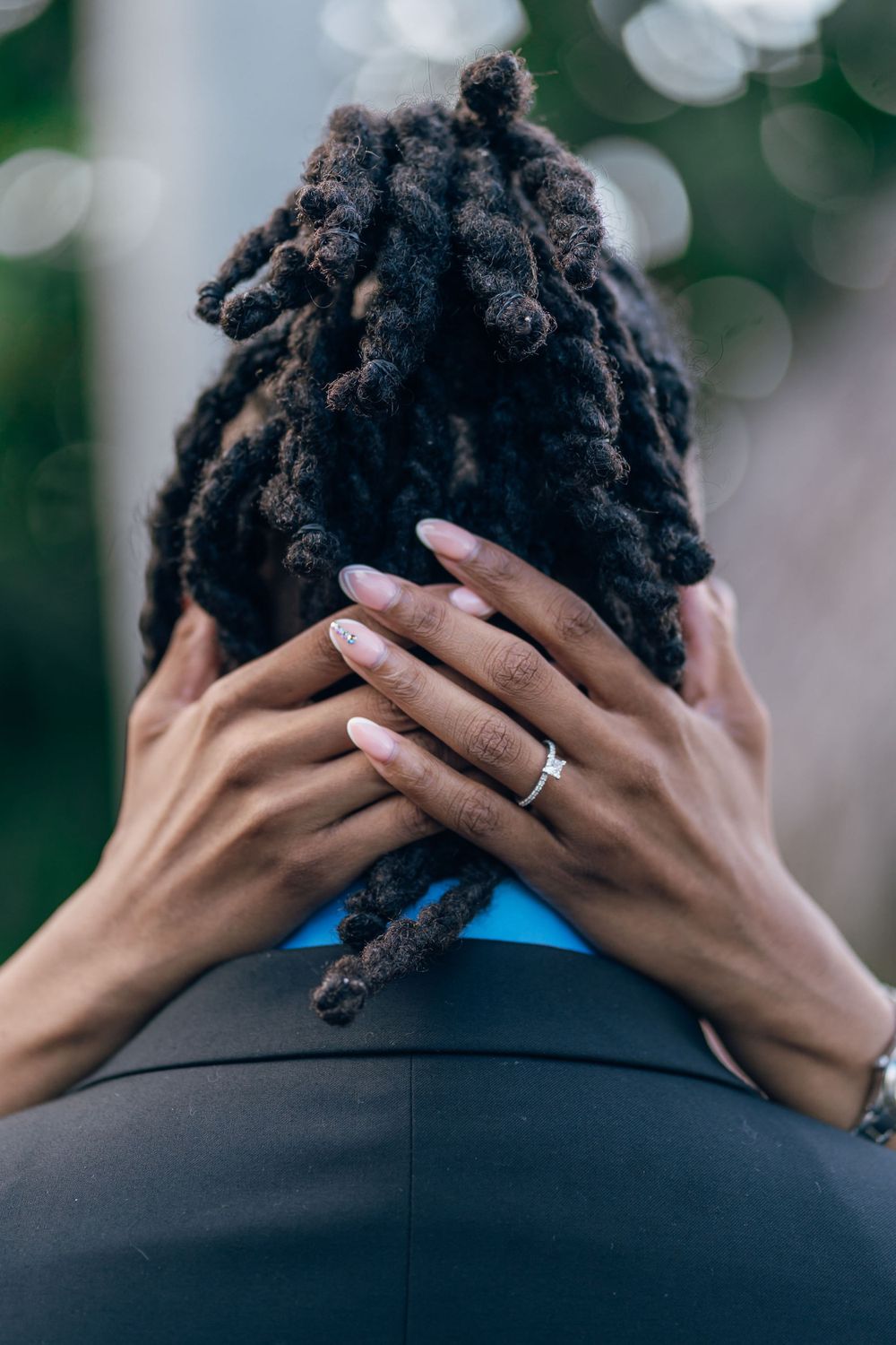 Hands wearing engagement ring rest on styled dreadlocks against blurred natural background.