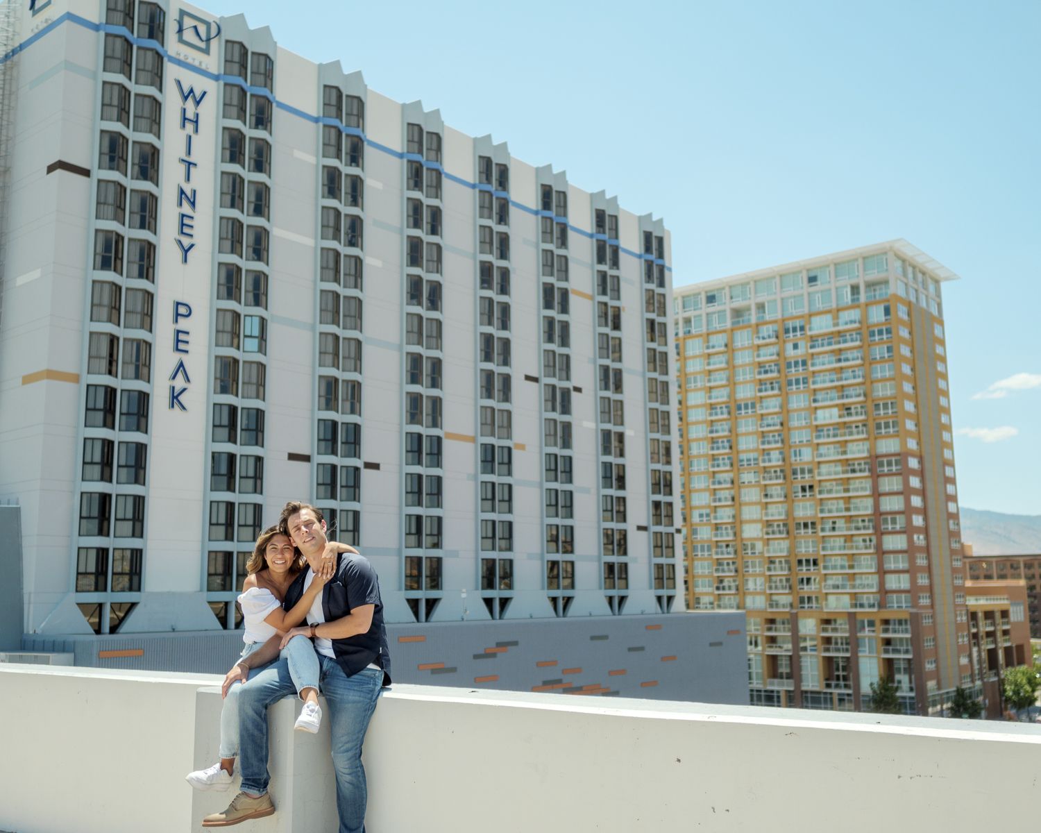 Two people sit together on an urban rooftop with tall residential buildings visible in the background.