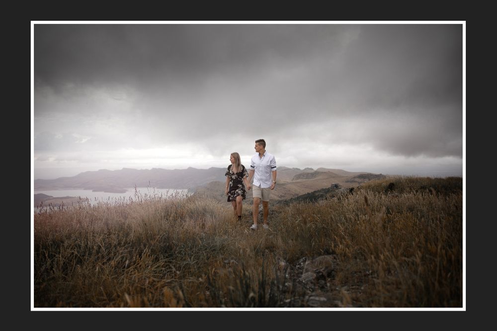 Couple standing on hilltop overlooking mountainous landscape under moody sky.