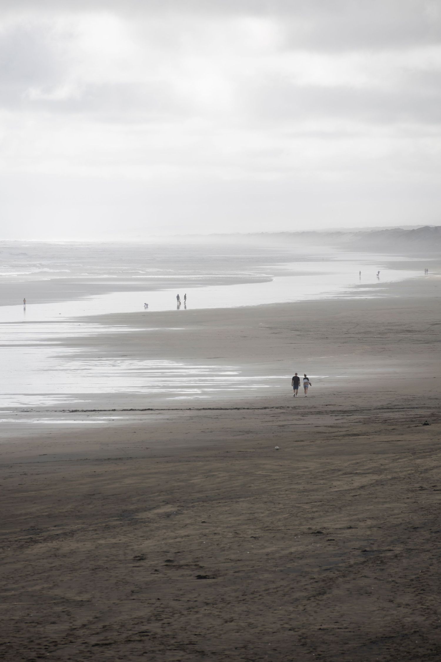 A cloudy beach scene with scattered people walking along the wet sand, fading into the misty distance.