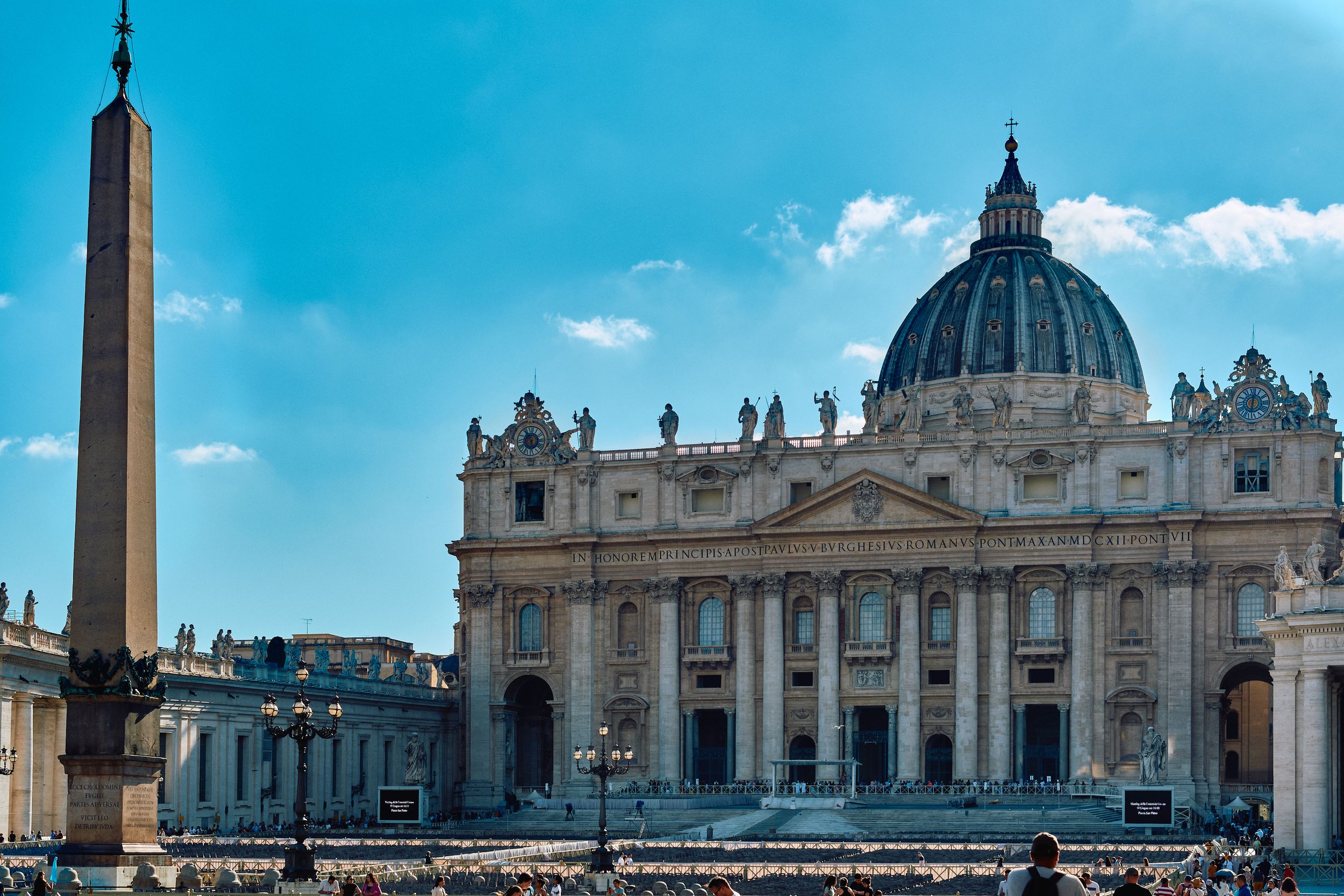 Saint Peter's Basilica facade and dome with Egyptian obelisk in foreground against vivid blue sky.