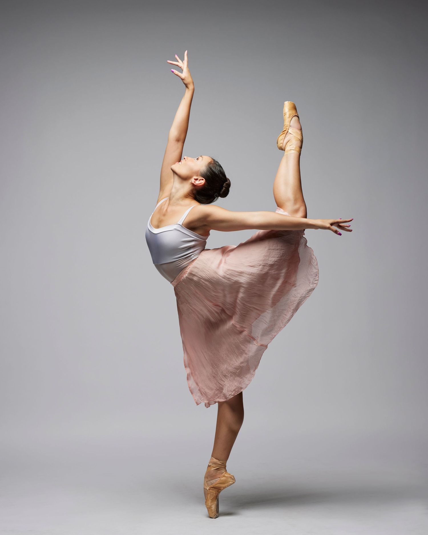 Ballet dancer in white top and pink skirt performing an arabesque pose against gray background.